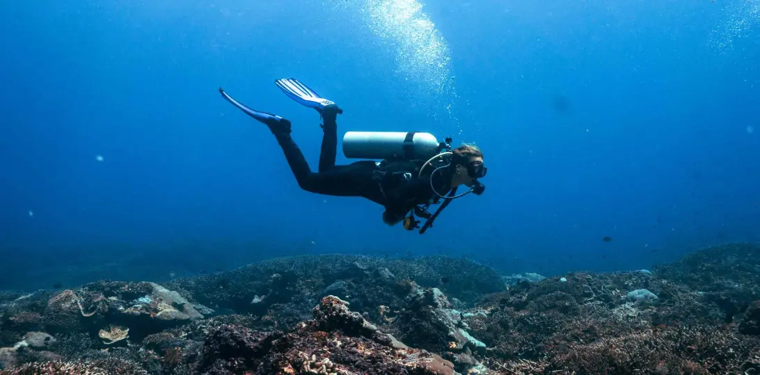 Taucherin mit Tauchgerät und Flossen schwimmt über Korallenriff bei Nusa Penida und atmet Luftblasen ins blaue Wasser.
