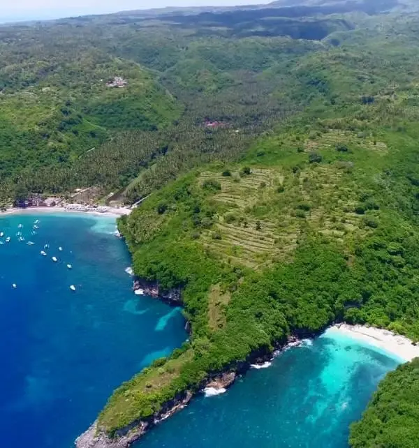 Aerial view of Nusa Penida showing turquoise bays, white-sand beaches, anchored boats and terraced green hills.
