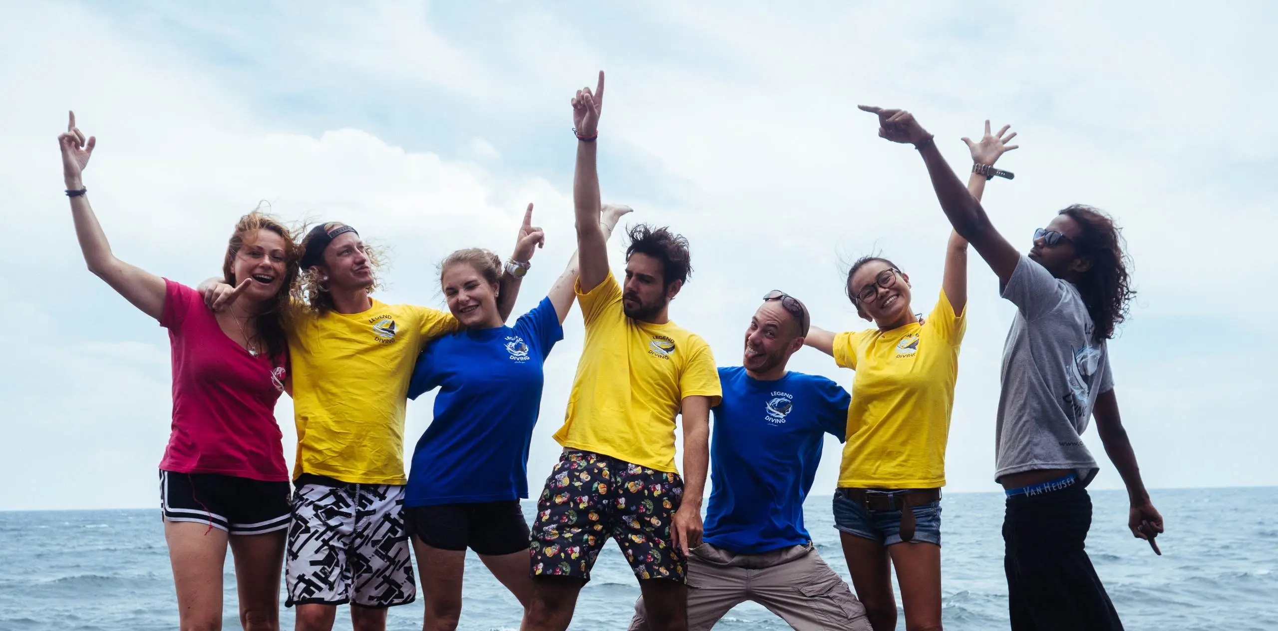 Seven people on Nusa Penida pose and cheer with arms raised, smiling against the ocean backdrop.