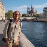 Mann mit Sonnenbrille und Rucksack lächelt am Ufer der Seine, im Hintergrund die Notre-Dame und eine Steinbrücke bei blauem Himmel.