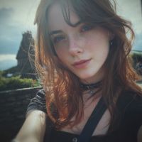 Junge Frau mit roten Haaren und leichten Sommersprossen, nahe Kamera lächelnd, wolkiger Himmel und Felsklippen im Hintergrund